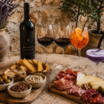 planche apéritives de charcuterie et fromages a droite, planche apéritifs provençales a gauche. derrière une bouteille de vin rouge beauté de nuit, deux verres de vin rouge, un cocktail orange et un cocktail violet